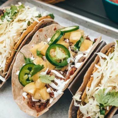 Three tacos in a steel rack on a tray, topped with cabbage, cilantro, pineapple and jalapeño, with lime wedges and a bowl of salsa