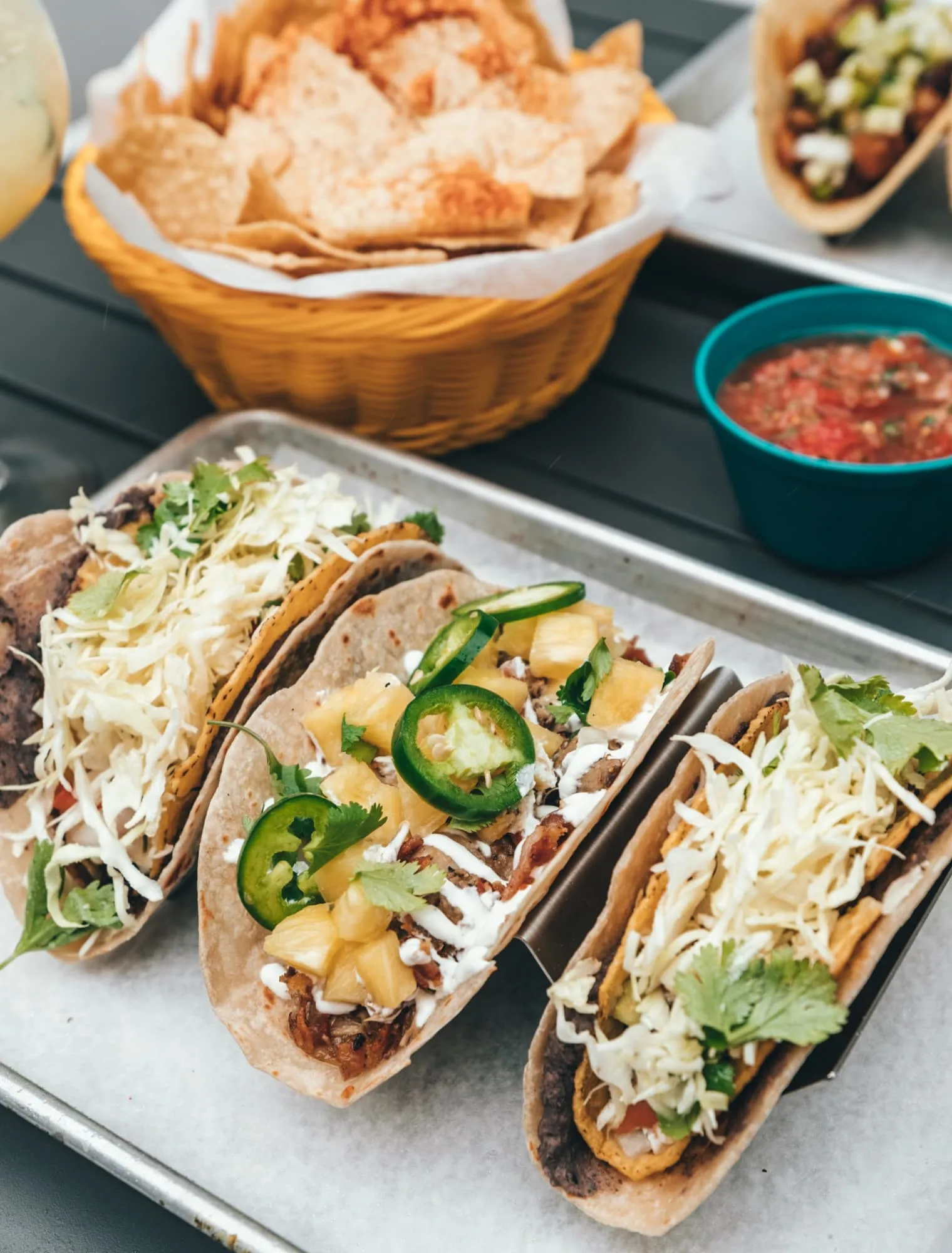 Tacos in a steel rack on a tray, topped with cabbage, cilantro, pineapple and jalapeño, with a basket of chips and a bowl of salsa behind